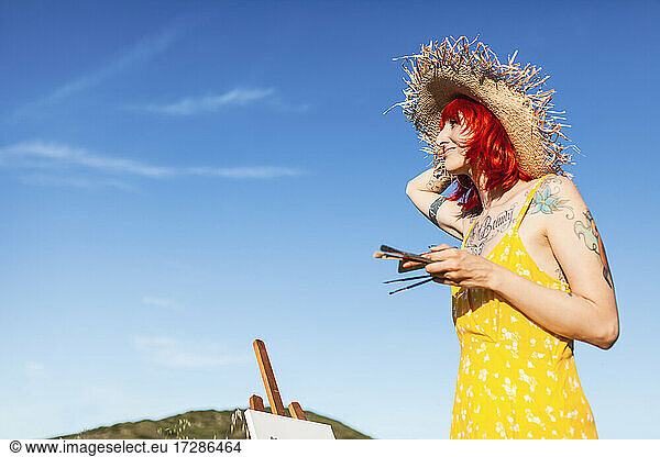 Tattooed female artist with paintbrushes looking away while holding sun hat