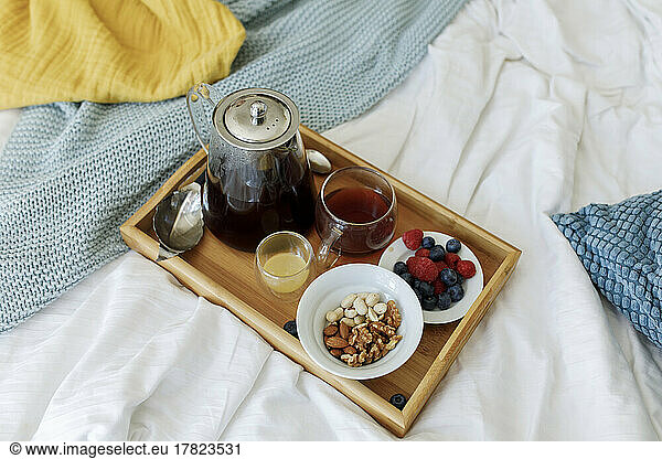 Tea pot in wooden tray with berries and nuts on bed