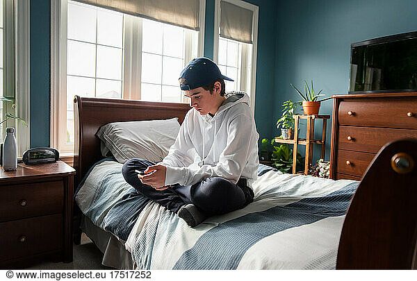 Teen boy in white hoodie on smartphone alone in his bedroom.