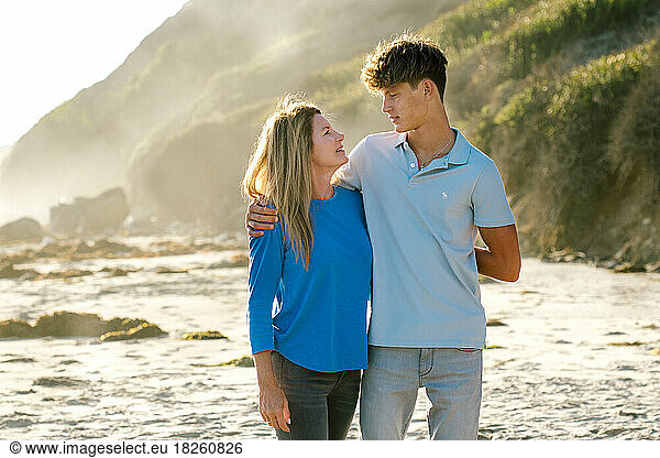 Teen Boy Looks Down At His Mother During An Embrace At The Beach