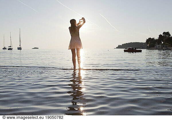 Teen girl frames up photo of sunset over lake