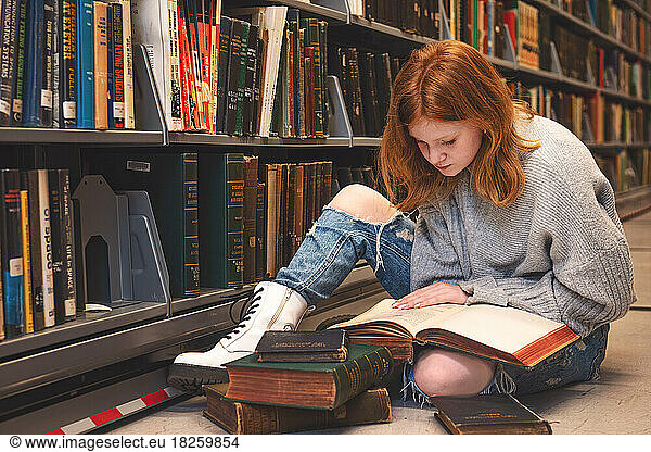Teen girl sitting on the floor reading in library.