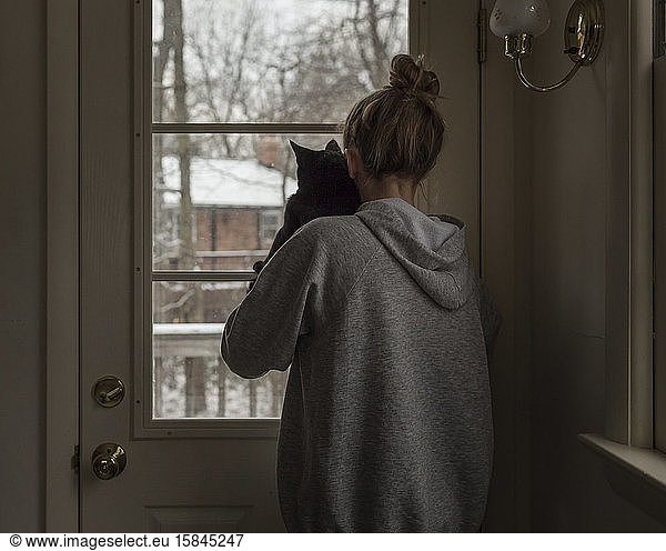 Teen girl wearing hoodie takes cat to look out window on snowy day