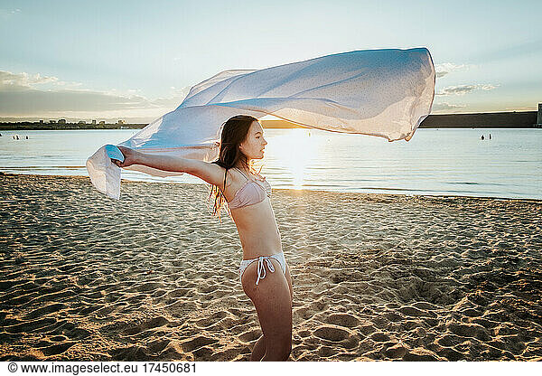 Teen girl wrapping sheet around her on the beach at sunset
