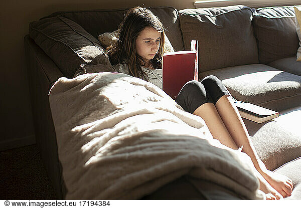 Teen girl 12-13 years old sits on couch and reads a book in the sun