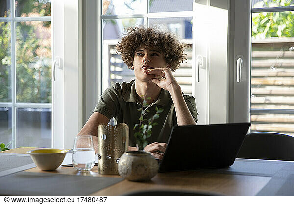 Teenage boy contemplating while sitting with laptop at home