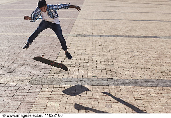 Teenage boy flipping skateboard on sunny cobblestone