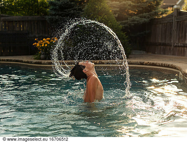 Teenage boy flipping water from his hair in an arc in a backyard pool.