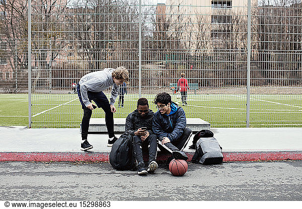 Teenage boy looking at friends using social media on sidewalk against soccer field in city