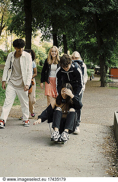 Teenage boy pushing cheerful girl sitting on skateboard while spending leisure time with friends in park
