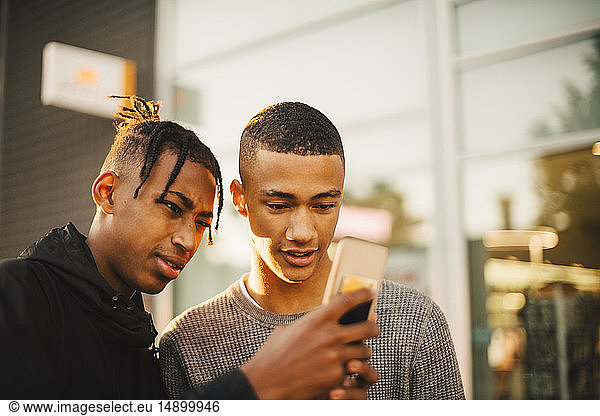 Teenage boy showing mobile phone to friend in city during sunset