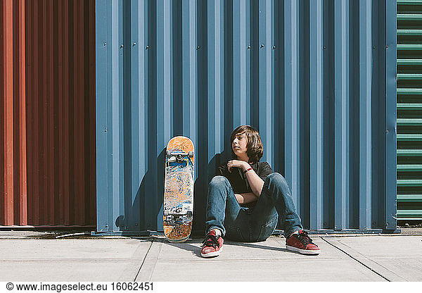 Teenage boy sitting with skateboard against warehouse wall