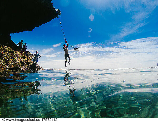 Teenage boy swings on rope across teal blue water in summer