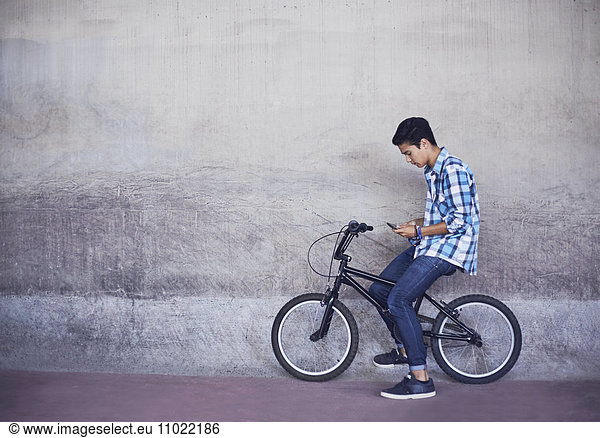 Teenage boy texting on BMX bicycle at wall