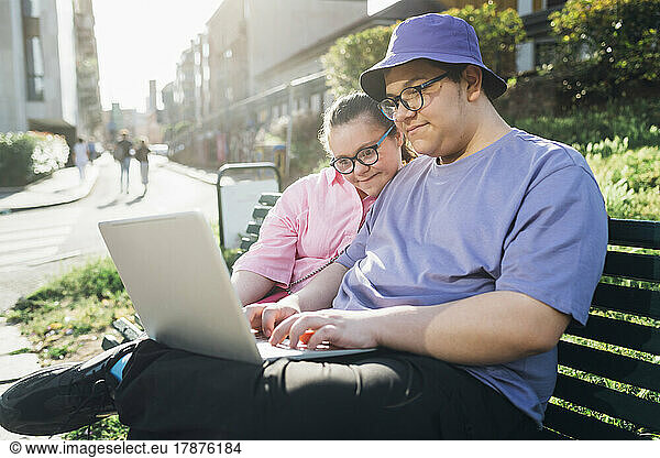 Teenage boy using laptop by sister with down syndrome on bench