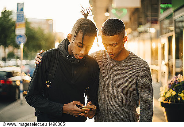 Teenage boy using mobile phone while standing by friend on street in city during sunset