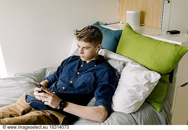 Teenage boy with in-ear headphones using smart phone while sitting on bed at home