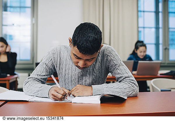 Teenage boy writing on book while studying at desk in high school