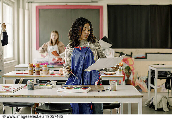 Teenage female student examining painting standing near desk during art class at high school