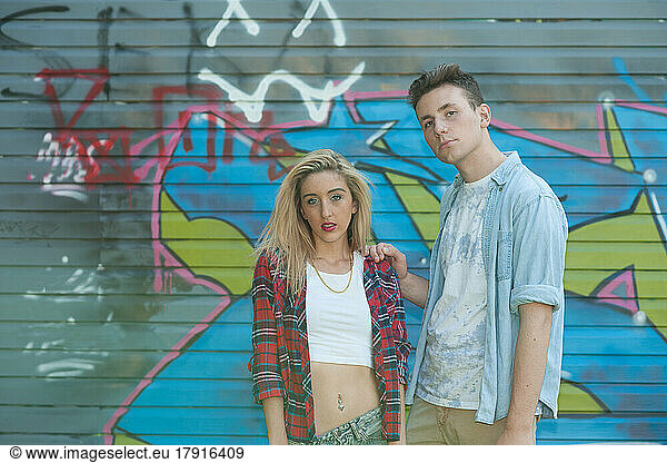 Teenage girl and boy standing in front of graffiti on wall.