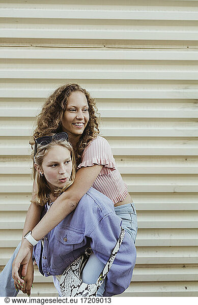 Teenage girl carrying female friend piggyback against metal wall