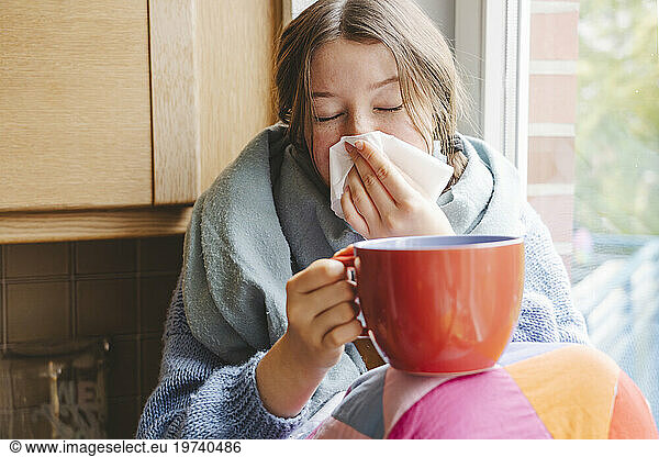 Teenage girl holding big coffee cup and cleaning nose with tissue at home