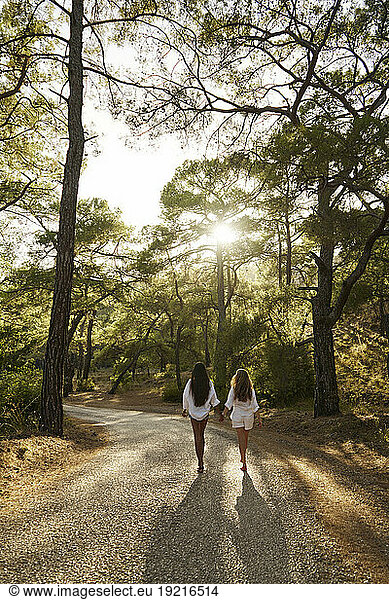 Teenage girl holding hands with mother walking on road in forest at sunset