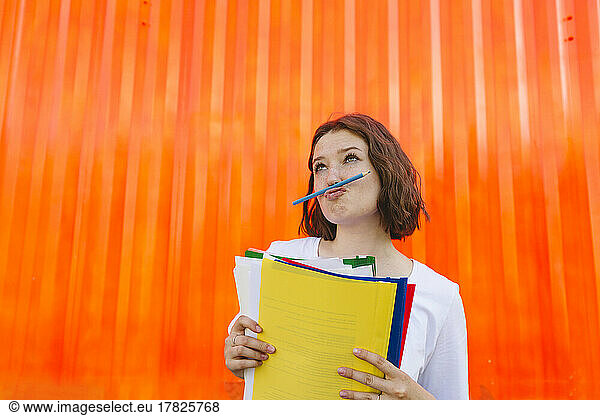 Teenage girl holding multi colored file folders puckering lips in front of orange cargo container
