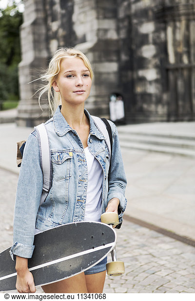 Teenage girl holding skateboard walking on cobbled street in city