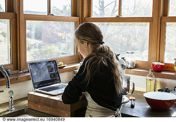 Teenage girl in a kitchen following a baking recipe on a laptop.