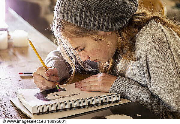 Teenage girl in a woolly hat drawing with a pencil on a sketchpad.