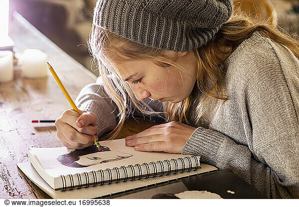 Teenage girl in a woolly hat drawing with a pencil on a sketchpad.
