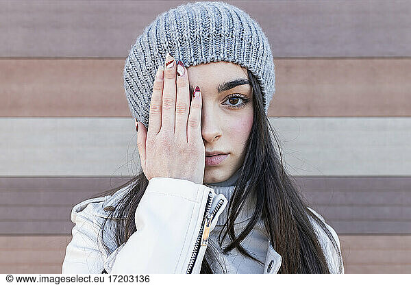 Teenage girl in knit hat covering her eyes with hands against wall