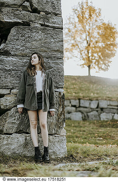 teenage girl in skirt stands against stone wall in fall