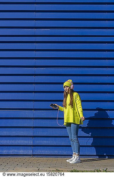 Teenage girl listening music with headphones and smartphone in front of blue background