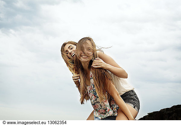 Teenage girl piggybacking female friend against sky