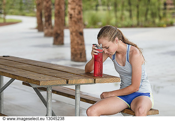 Teenage girl resting head on water bottle while sitting on bench