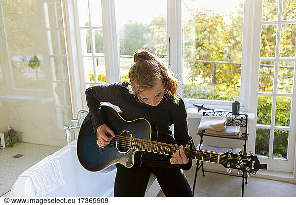 Teenage girl seated on the edge of a bathtub  playing accoustic guitar.