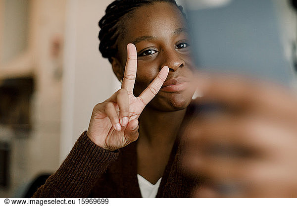 Teenage girl showing peace sign while taking selfie through smart phone at home