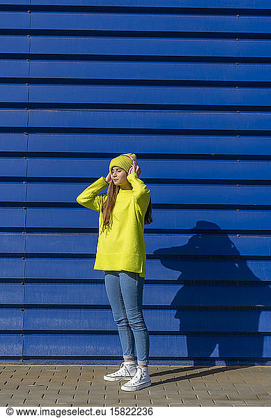 Teenage girl standping in front of blue background listening music with headphones