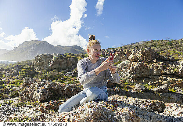 Teenage girl taking pictures with her smart phone  sitting on the rocks on a beach