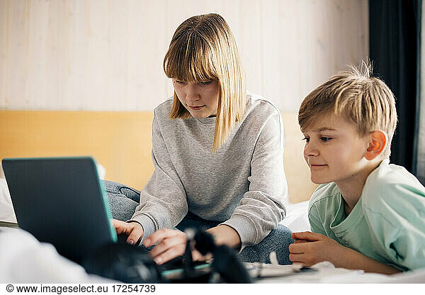 Teenage girl using laptop while sitting by brother on bed at home