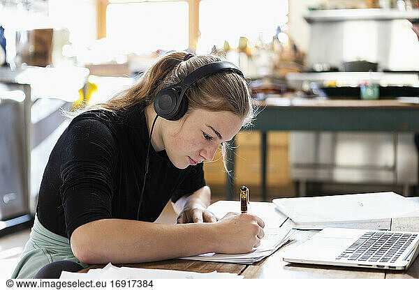 Teenage girl wearing headphones using a laptop and writing in a notebook