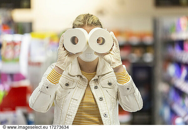 Teenage girl wearing protectice mask and gloves holding looking through holes of toilet rolls at supermarket