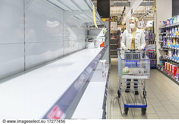 Teenage girl wearing protectice mask and gloves looking at empty shelves at supermarket