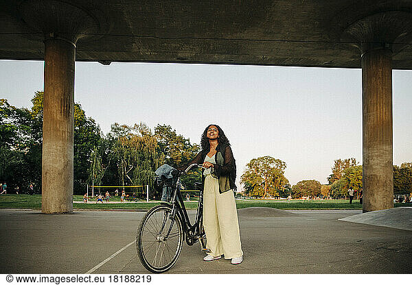 Teenage girl with bicycle standing below bridge