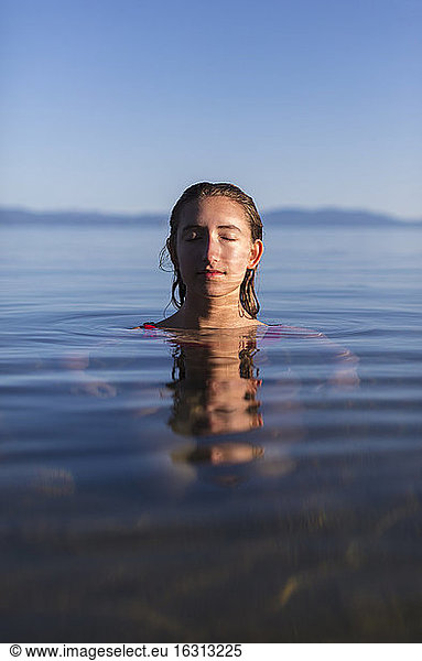 Teenage girl with eyes closed  head and shoulders above the calm water of a lake at dawn