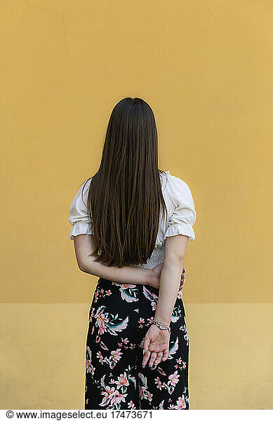 Teenage girl with hands behind back standing against yellow background