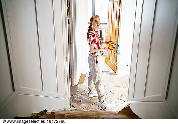Teenage girl with paint roller standing near door at home