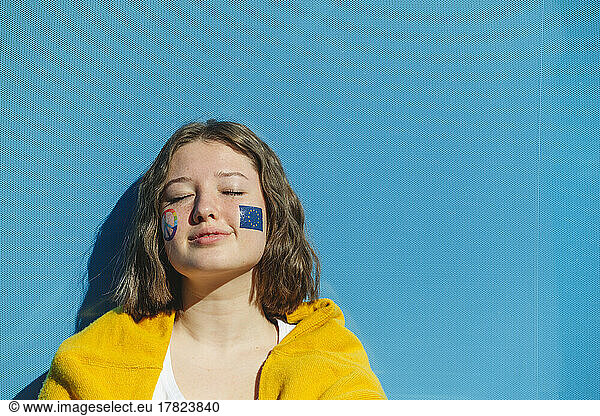 Teenage girl with peace symbol and European Union paint on cheeks in front of blue wall